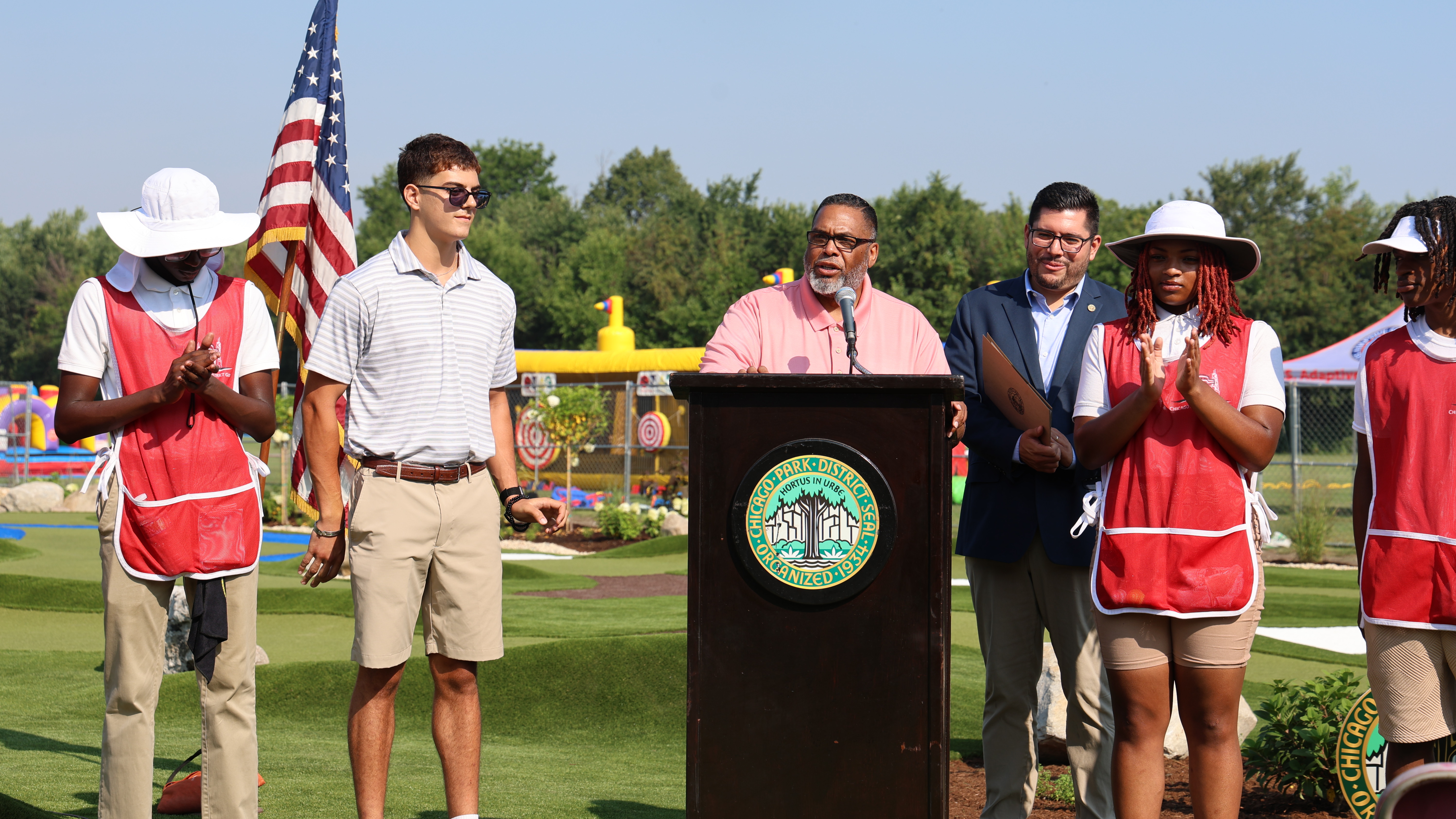 A speaker at a podium addresses attendees at a park district event.  An American flag and mini golf course are in the background.